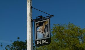 Picture of the painted pub sign on a white post (that could do with a repaint) against a glorious blue sky. The composition of the painting is taken from one of the original signs from years ago - of a bell clanging in a belfry. The words The Bell are written underneath in light writing with a dark green background. The sign is located on the triangle of grass in the car park.
